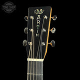 Close-up of a guitar headstock with the brand name MARTIN vertically carved in gold on a black background, with six tuning pegs and six strings visible. A Tone Shop watermark is in the top-left corner.