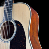 Close-up of an acoustic guitar showing the natural wood soundboard, dark pickguard, black fretboard, and rich reddish-brown side panel against a black background.
