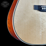 Close-up of an acoustic guitar body showing the natural maple wood top, the reddish-brown edge, and part of the black bridge against a black background.