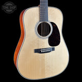 A close-up studio shot of an acoustic guitar body against a solid black background, featuring a natural wood top, dark reddish-brown sides, a black pickguard, and a white brand logo inlay on the lower bout.