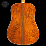 Close-up of the back of an acoustic guitar showing a rich, warm orange-brown wood grain finish with a white edge trim, set against a solid black background. A Tone Shop watermark is visible in the top-left corner.