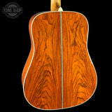 Close-up of the back of an acoustic guitar showing a rich, warm reddish-brown wood grain finish with a white binding edge, set against a solid black background.