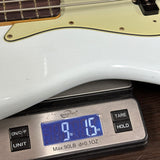 A close-up of a white electric bass guitar body resting on a digital scale displaying 9.15 lbs, with part of the neck and a cream pickguard visible.