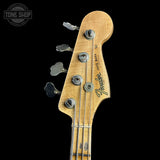 Close-up of a Fender Jazz Bass headstock with a natural wood finish, four silver tuning pegs, and the Fender logo, shown against a solid black background.