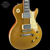 A classic electric guitar with a distressed gold finish, featuring a cream pickguard, gold hardware, and black humbucker pickups, shown against a solid black background.