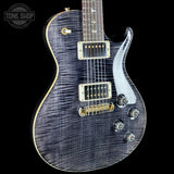 A close-up of an electric guitar body with a dark blue-gray flamed maple finish, gold hardware, and a white pickguard, shown against a black background with a Tone Shop watermark in the top left.