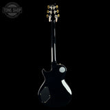 A black electric guitar shown from the back against a solid black background, featuring gold tuning pegs and a white circular detail on the body.