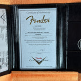 A black leather Fender instrument authenticity certificate insert displayed open, showing a silver certificate with the Fender logo and model details, photographed against an orange textured background.