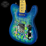 A blue electric guitar with a vibrant floral pattern in pink, green, and yellow on the body, featuring a maple neck and chrome hardware, shown against a black background with a Tone Shop watermark.