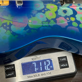 A close-up of a blue electric guitar body with a colorful floral pattern, resting on a digital scale displaying 7.12 oz.