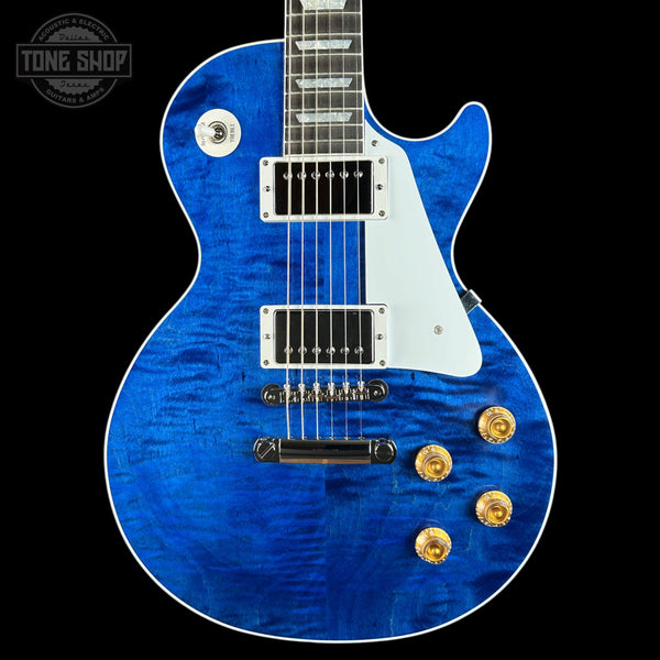 A blue flamed maple top electric guitar with a white pickguard and gold hardware, shown against a solid black background. A Tone Shop logo is visible in the top-left corner.