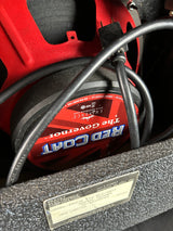 A close-up of a Red Coat 1100X speaker inside a custom car trunk, showing the red and black speaker cone, black power cord, and a silver metal plate in the trunk lid.