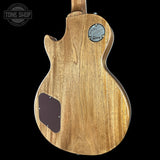 The back of an electric guitar with a natural wood finish, featuring a circular metal Gibson logo badge and a dark brown pickguard, shown against a solid black background.