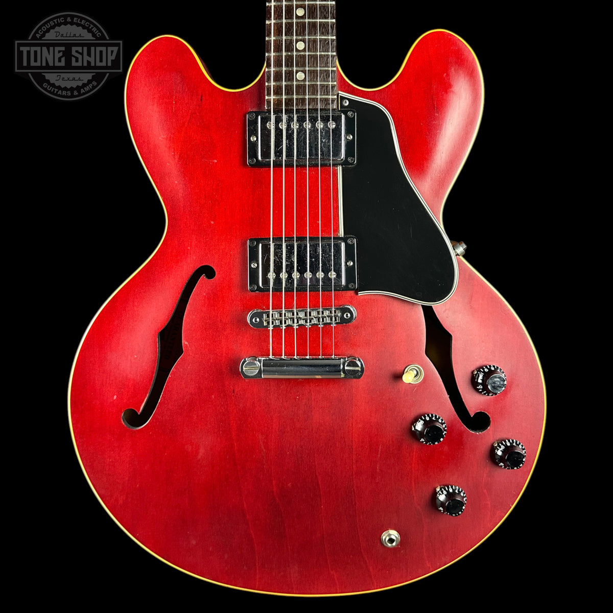 A red electric guitar with a black pickguard, dual humbucker pickups, and chrome hardware, shown against a black background with a Tone Shop watermark in the top left corner.
