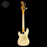 A Fender electric bass guitar shown from the back, featuring a cream-colored body with heavy distressed scuffing, an orange maple neck, and gold tuning pegs, set against a solid black background.