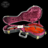An open black guitar case with bright pink velvet lining, holding a double-cutaway mandolin guitar. The mandolin has a glossy orange-brown body, a white headstock, and gold hardware.
