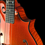 Close-up of a mandolin with a glittery orange-red finish and black binding, showing part of the fretboard and f-holes against a black background.
