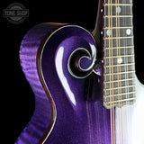 Close-up of a purple glitter electric guitar body with a scroll cutout and part of the fretboard visible, photographed against a black background. A Tone Shop watermark appears in the upper left.