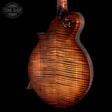 Close-up of the back of a mandolin with a rich, figured wood grain finish in warm brown and amber tones, set against a solid black background with a Tone Shop watermark in the top left corner.
