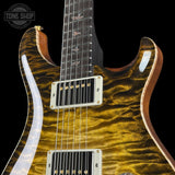 Close-up of a double-cutaway electric guitar body with a glossy flamed maple top in a metallic gold finish, featuring white hardware and a dark fretboard, shown against a black background.