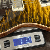 Close-up of a guitar body with a glossy wavy gold and amber finish resting on a digital scale displaying 7.32 oz.