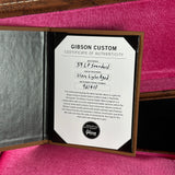 An open Gibson Custom presentation case showing a Certificate of Authenticity insert for a 59 LP Stranded ultra light aged instrument, placed on a bright pink velvet surface.