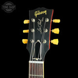Close-up of the headstock of a Gibson Les Paul Model electric guitar, featuring a black headstock with yellow tuning pegs, gold Gibson script, and a red edge outline, set against a solid black background.