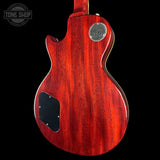 The back of an electric guitar with a glossy reddish-orange wood finish, featuring a circular metal 'Gibson' logo badge and a black pickguard, shown against a solid black background.