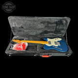 A blue electric guitar with a cream pickguard and yellow maple neck, displayed inside an open black hard case with gray foam lining. A red accessory in a plastic bag is tucked under the guitar.