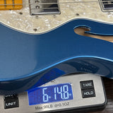 A close-up of a blue electric guitar body resting on a digital scale displaying 6.140 oz, with part of the cream pickguard and pickups visible above.