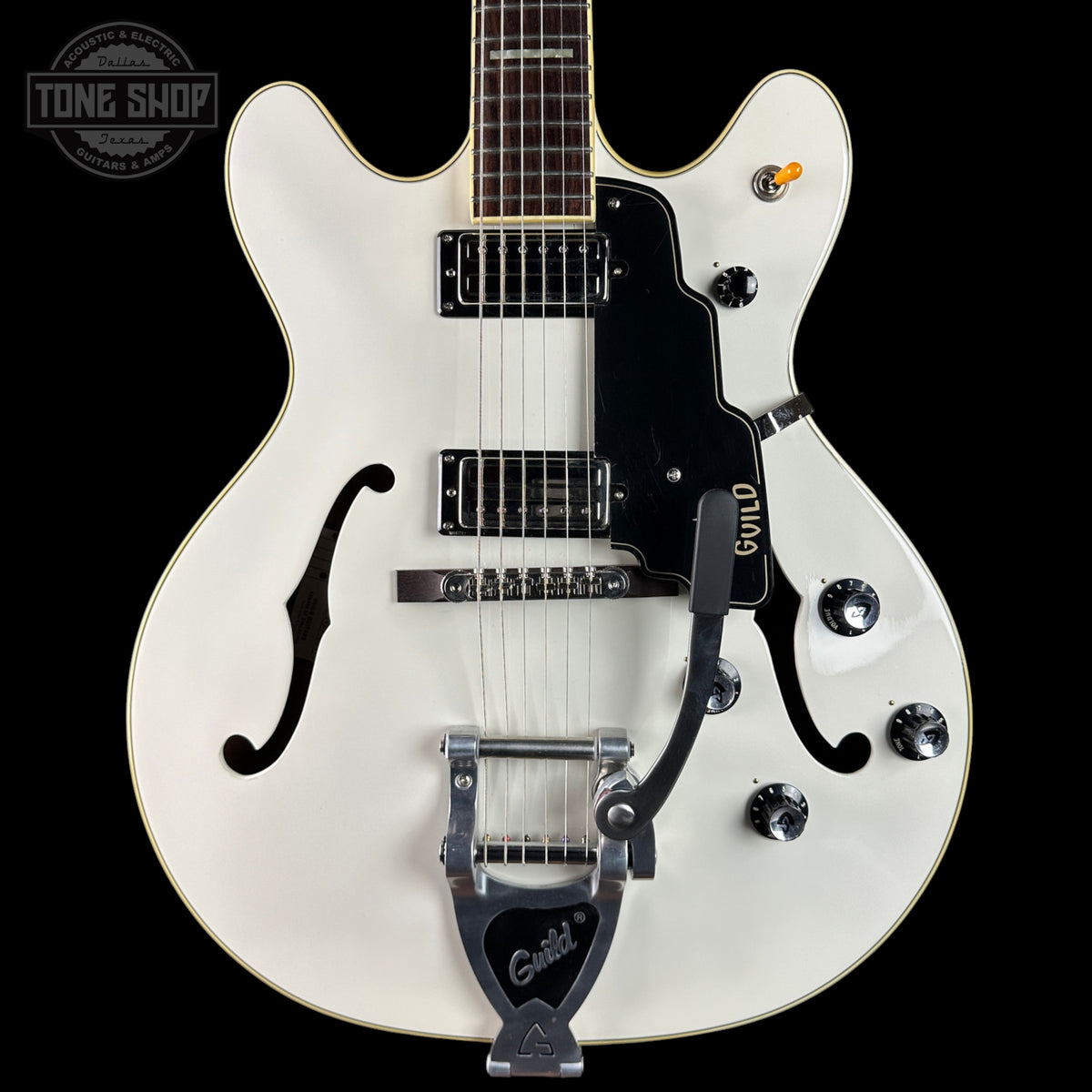 White electric guitar with a black pickguard on a black background, featuring the Guild logo.