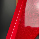Close-up of a glossy red metallic surface with fine glitter/sparkle texture and a bright reflective highlight against a dark gray background.