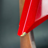 Extreme close-up of a glossy red painted metal surface, likely a vehicle fender or bumper edge, showing a small gold-toned paint chip or bare metal area near the tip against a blurred dark gray and brown background.