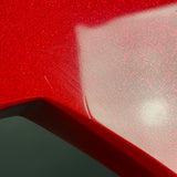 Close-up of a glossy red car body panel with fine glitter/sparkle texture and a bright reflection, showing a small scratch along the edge.