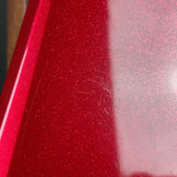 Close-up of a glossy red glitter surface with fine pinkish-red sparkle, showing a vertical highlight and a few small scratches on the finish.