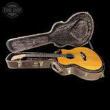 An acoustic guitar with a natural wood finish resting inside an open brown hard case on a black background. A Tone Shop watermark is visible in the top left corner.