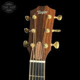 Close-up of the headstock of a Taylor acoustic guitar, showing the brand name in white script on a dark wood background, six gold tuning pegs, and strings extending down the fretboard against a black background.