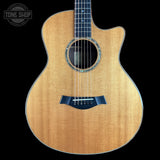 A front-facing acoustic guitar with a natural wood finish and a dark navy blue bridge, shown against a solid black background with a Tone Shop watermark in the top-left corner.