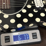 A close-up of a black electric guitar body with large cream polka dots resting on a digital scale displaying 6.77 oz.