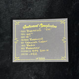 A gray rectangular specification card with yellow printed text and border, placed on a black textured surface. The card reads 'Instrument Specification' and lists details such as pickups, profile, fingerboard, body, and other guitar components.