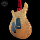 Close-up of the back of an electric guitar body with a natural wood finish, black pickguard, and red neck visible against a black background. A Tone Shop watermark appears in the upper left.