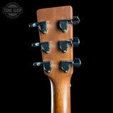 Close-up of the headstock of a six-string acoustic guitar, showing the six black tuning pegs and a warm brown wood finish, set against a solid black background with a Tone Shop watermark in the top left corner.