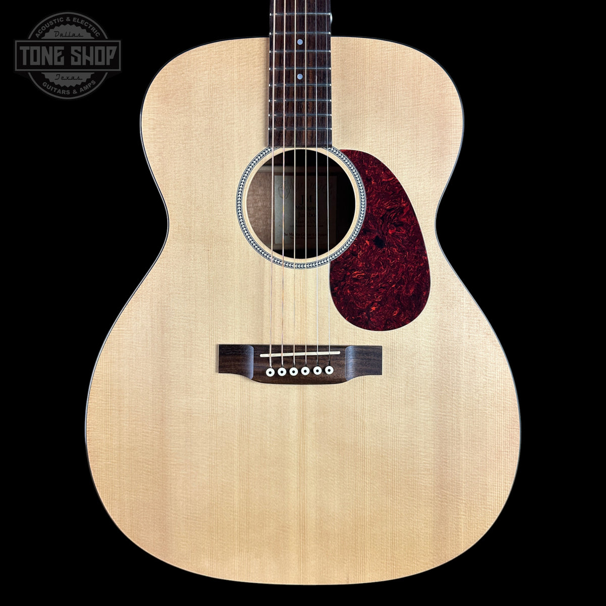 An acoustic guitar with a natural wood top and a dark red tortoiseshell pickguard, shown against a solid black background with a Tone Shop logo in the upper left corner.
