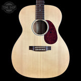 An acoustic guitar with a natural wood top and a dark red tortoiseshell pickguard, shown against a solid black background with a Tone Shop logo in the upper left corner.