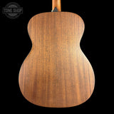 Close-up of the back of an acoustic guitar showing a warm brown wood grain finish, set against a solid black background with a Tone Shop watermark in the top left corner.