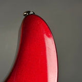 Close-up of a glossy red curved object, likely part of a guitar body or similar instrument, with a small silver metal detail at the top against a plain gray background.