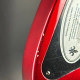 Close-up of a red electric guitar body showing the edge of the finish, a black pickguard with visible scratches, and part of the silver manufacturer plate with text and a fleur-de-lis logo.