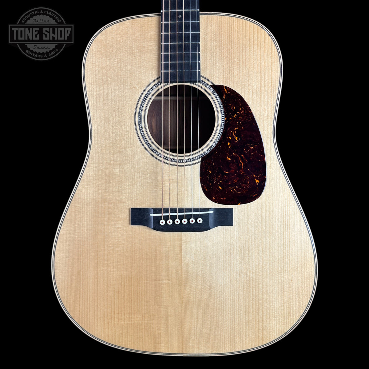 Front view of an acoustic guitar with a natural wood finish, black neck, and dark tortoiseshell pickguard on a black background. A Tone Shop logo is visible in the top-left corner.