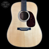 Front view of an acoustic guitar with a natural wood finish, black neck, and dark tortoiseshell pickguard on a black background. A Tone Shop logo is visible in the top-left corner.