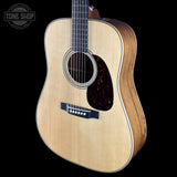 A close-up studio shot of an acoustic guitar against a solid black background, featuring a natural wood body, dark brown pickguard, and black bridge. A Tone Shop logo is visible in the top-left corner.